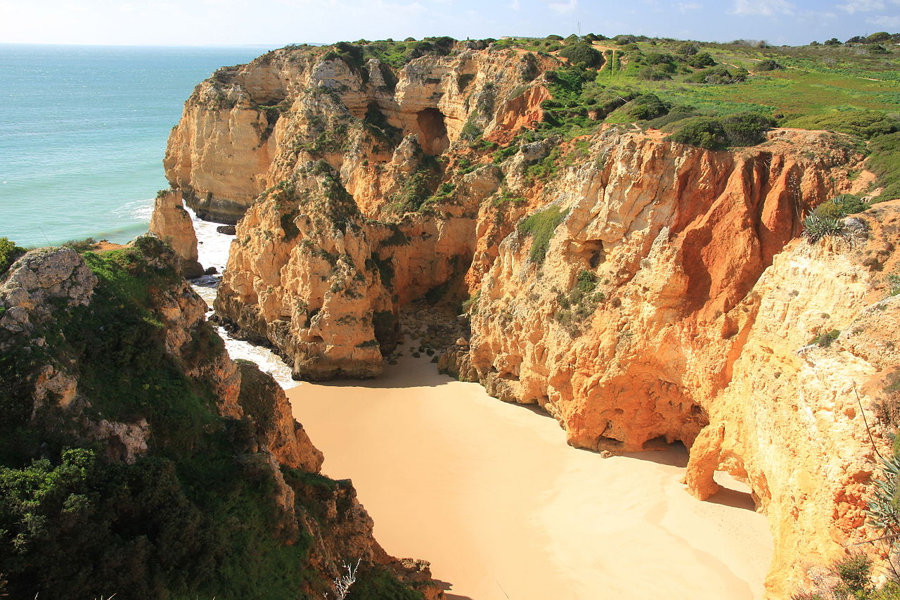 Portugalia - Algarve - klify w pobliżu Lagos” /><figcaption>Zdjęcie: muffinn z Worcester w Wielkiej Brytanii, CC BY 2.0, za pośrednictwem Wikimedia Commons.</figcaption></figure>
<h2>Praia Dona Ana – plaża idealna na pocztówkę</h2>
<p>Zacznę od plaży, która sprawiła, że zakochałam się w Lagos. Praia Dona Ana niezmiennie zaliczana jest do najpiękniejszych plaż na świecie i po zobaczeniu jej na własne oczy całkowicie rozumiem dlaczego. Otoczona spektakularnymi klifami w kolorze ochry i złotego wapienia, plaża charakteryzuje się krystalicznie czystą turkusową wodą i miękkim złotym piaskiem, który wydaje się być snem.</p>
<p>Przybyłem wcześnie rano, około 8:30 i miałem to miejsce prawie dla siebie. Światło o tej porze jest magiczne, rzuca ciepły złoty blask na formacje skalne. Ze szczytu klifu na plażę prowadzą drewniane schody, a widok, jaki schodzi się w dół, zapiera dech w piersiach.</p>
<p><strong>Moja rada:</strong> Przyjdź wcześnie lub późno w ciągu dnia. Latem do południa plaża szybko się zapełnia. Można wypożyczyć leżaki, ale wolałam znaleźć własne miejsce w pobliżu skał po wschodniej stronie, gdzie po południu jest więcej cienia.</p>
<h2>Praia do Camilo – ukryty klejnot</h2>
<p>W odległości krótkiego spaceru od Dona Ana Praia do Camilo to mniejsza, bardziej kameralna plaża schowana pomiędzy wysokimi klifami. Dojdziesz do niego, schodząc po stromych drewnianych schodach liczących około 200 stopni wykutych w ścianie klifu. Samo zejście jest przeżyciem, a za każdym zakrętem otwierają się panoramiczne widoki na wybrzeże.</p>
<p>Plaża jest podzielona na dwie małe zatoczki połączone naturalnym tunelem wykutym w skale przez wieki fal. Pływanie tym tunelem było jak wejście do sekretnego świata. Woda tutaj jest niezwykle spokojna i czysta, dzięki czemu idealnie nadaje się do snorkelingu wzdłuż skalnych ścian, gdzie można obserwować małe ryby i ukwiały.</p>
<p><strong>Uwaga:</strong> ponieważ plaża jest mała, szybko się zapełnia. W szczycie sezonu polecam przyjazd przed godziną 9:00. Pamiętaj też, że 200 schodów w dół oznacza 200 schodów z powrotem w upale.</p>
<h2>Ponta da Piedade – klejnot koronny Algarve</h2>
<p>Ponta da Piedade nie jest technicznie plażą, ale oszałamiającym cyplem z rzeźbionymi złotymi klifami, kominami, ukrytymi grotami i łukami wznoszącymi się do 20 metrów nad Atlantykiem. Jest to bez przesady jedna z najbardziej spektakularnych formacji naturalnych, jakie widziałem na świecie.</p>
<p>Szedłem tutaj z centrum Lagos ścieżką na szczycie klifu, co zajmuje około 40 minut i oferuje coraz bardziej zapierające dech w piersiach widoki w miarę zbliżania się do punktu. Znajdują się tam kamienne schody schodzące na małą platformę na poziomie wody, skąd można obserwować fale rozbijające się o formacje skalne i zaglądać do jaskiń lśniących niesamowitym odcieniem turkusu.</p>
<p>Jednak prawdziwą magią jest oglądanie Ponta da Piedade z wody. Wybrałem się na wycieczkę łodzią z przystani w Lagos i była to najlepsza aktywność, jaką zrobiłem podczas całego mojego pobytu w Portugalii. Więcej na ten temat poniżej.</p>
<h2>Meia Praia – bezkresny odcinek</h2>
<p>Po dniach zwiedzania intymnych zatoczek otoczonych klifami, Meia Praia była odświeżającą odmianą. Jest to masywna, szeroko otwarta plaża rozciągająca się około 4 kilometrów wzdłuż wschodniej strony zatoki Lagos. To taka plaża, po której można spacerować pół godziny i wciąż mieć przed sobą piasek.</p>
<p>Meia Praia jest idealna na długie dni na plaży. Jest dużo miejsca nawet w szczycie lata, a plaża ma dobre połączenie obsługiwanych obszarów z leżakami i restauracjami, a także dziksze, bardziej puste odcinki w kierunku wschodniego krańca. Woda jest tu nieco cieplejsza, ponieważ zatoka jest bardziej osłonięta, i jest to również najlepsze miejsce w Lagos do uprawiania sportów wodnych, takich jak kitesurfing i paddleboarding na stojąco.</p>
<p><strong>Jak się tam dostać:</strong> Z centrum Lagos można dojść pieszo przez kładkę w pobliżu przystani, około 15 minut. Można też skorzystać z małego promu, który przepływa przez rzekę ze starego miasta.</p>
<h2>Praia dos Estudantes – plaża miejska z niespodzianką</h2>
<p>W samym sercu Lagos, schowana pod murami starego miasta, Praia dos Estudantes to plaża, do której najłatwiej dotrzeć. To, co czyni ją wyjątkową, to most z czasów rzymskich, który wygina się łukiem nad skałami, łącząc plażę z drugą mniejszą zatoczką. To piękne i historyczne miejsce, które sprawia wrażenie niemal teatralnego.</p>
<p>Uwielbiałem przychodzić tu późnym popołudniem po zwiedzaniu starego miasta. Plaża pięknie łapie złote wieczorne światło, a w odległości dwóch minut spacerem znajduje się kilka znakomitych restauracji i barów. Jest mała i robi się tłoczno, ale atmosfera jest żywa i zabawna.</p>
<h2>Porto de Mos – lokalny faworyt</h2>
<p>Nieco dalej od centrum Porto de Mos to szeroka, piękna plaża otoczona zielonymi klifami. Ma bardziej zrelaksowaną, lokalną atmosferę w porównaniu do słynnych plaż położonych bliżej miasta. Klify są tu oszałamiające, z warstwami pomarańczowego, żółtego i białego wapienia świecącymi w popołudniowym słońcu.</p>
<p>Jest tu kilka doskonałych restauracji przy plaży, a fale są zwykle nieco większe niż w osłoniętych zatoczkach, co czyni to miejsce fajnym do uprawiania bodyboardingu. Spędziłem tu całe popołudnie i był to jeden z najbardziej relaksujących dni mojej podróży.</p>
<p><strong>Dojazd:</strong> Porto de Mos znajduje się około 25 minut spacerem od centrum Lagos lub 5 minut jazdy samochodem. Na szczycie klifu znajduje się bezpłatny parking.</p>
<h2>Groty i jaskinie morskie – obowiązkowy rejs łodzią</h2>
<p>Nie można odwiedzić Lagos bez rejsu łodzią po słynnych grotach i jaskiniach morskich wzdłuż wybrzeża. Cały odcinek między Ponta da Piedade a Praia Dona Ana jest usiany jaskiniami, łukami i tunelami, do których można dostać się tylko od strony wody.</p>
<p>Zarezerwowałem wycieczkę w małej grupie tradycyjną łodzią rybacką z przystani w Lagos i trwała ona około 75 minut. Jechaliśmy wzdłuż wybrzeża, zanurzając się w wąskich jaskiniach, gdzie woda lśniła niebiesko-zielonym blaskiem, mijając naturalne łuki i zatrzymując się na ukrytych plażach, do których można dotrzeć tylko łodzią. Kapitan znał każdą jaskinię i idealnie zgrał podejście z falami.</p>
<h3>Wskazówki dotyczące rezerwacji wycieczek łodzią</h3>
<ul>
<li><strong>Małe łodzie są lepsze:</strong> mniejsze, tradycyjne łodzie mogą wpływać do jaskiń, do których nie mogą dotrzeć większe jednostki. Poszukaj łodzi przewożących maksymalnie 10–15 osób.</li>
<li><strong>Poranne wycieczki zapewniają spokojniejsze morze</strong> i lepsze światło w jaskiniach. Poszedłem o 10 rano i było idealnie.</li>
<li><strong>Zarezerwuj z wyprzedzeniem</strong> w lipcu i sierpniu. Wycieczki wyprzedają się, szczególnie te poranne.</li>
<li><strong>Za standardową wycieczkę po grocie można spodziewać się</strong> około 20–30 euro od osoby.</li>
<li><strong>Wycieczki kajakiem</strong> są również dostępne, jeśli chcesz bardziej aktywnego doświadczenia. Trwają dłużej, ale oferują jeszcze bardziej intymną perspektywę.</li>
</ul>
<h2>Praktyczne informacje dotyczące zwiedzania Lagos</h2>
<h3>Jak dojechać do Lagos</h3>
<p><strong>Z Lizbony:</strong> Lagos leży około 290 km na południe od Lizbony. Można dojechać bezpośrednim pociągiem ze stacji Estacao do Oriente lub Entrecampos w Lizbonie, podróż zajmuje około 3,5-4 godzin i kosztuje około 22-30 euro. Alternatywnie autobus Rede Expressos jedzie około 3,5 godziny i jest często tańszy.</p>
<p><strong>Z lotniska w Faro:</strong> Faro to główne lotnisko w Algarve, około 90 km na wschód od Lagos. Można dojechać pociągiem ze stacji Faro (około 1,5-2 godziny z przesiadką w Tunes) lub dojechać w około godzinę autostradą A22. Wielu gości wynajmuje samochód na lotnisku w Faro, co zapewnia największą elastyczność w zwiedzaniu regionu.</p>
<p><strong>Poruszanie się po Lagos:</strong> Do samego miasta można łatwo dojść pieszo. Do wszystkich plaż, o których wspomniałem, można dotrzeć pieszo, choć niektóre spacery zajmują 25-40 minut. Istnieje również lokalny autobus, a na dłuższych dystansach taksówki są niedrogie.</p>
<h3>Najlepszy czas na wizytę</h3>
<p><strong>Od maja do czerwca</strong> to moja najlepsza rekomendacja. Pogoda jest ciepła i słoneczna (około 22-26 stopni Celsjusza), plaże nie są jeszcze zapełnione letnimi tłumami, woda jest orzeźwiająca, a ceny są niższe niż w sezonie.</p>
<p><strong>Lipiec i sierpień</strong> przynoszą najcieplejszą pogodę i najwyższe temperatury wody, ale także największe tłumy i najwyższe ceny. Jeśli wybierasz się tu w szczycie lata, przybądź na plaże wcześniej.</p>
<p><strong>Wrzesień i październik</strong> to doskonała alternatywa. Latem tłumy przerzedzają się, ocean jest najcieplejszy po miesiącach słonecznych, a pogoda utrzymuje się piękna aż do października.</p>
<h3>Wskazówki dotyczące plaży</h3>
<ul>
<li><strong>Atlantyk jest zimny.</strong> Nawet w sierpniu temperatura wody oscyluje wokół 19–21 stopni Celsjusza. Na miejscu jest orzeźwiająco, ale przygotuj się na początkowy szok.</li>
<li><strong>Noś ochronę przeciwsłoneczną.</strong> Słońce w Algarve jest mocne, a odbicie od klifów i wody je wzmacnia. Pierwszego dnia poparzyłem się słońcem, mimo że myślałem, że zachowałem ostrożność.</li>
<li><strong>Zabierz ze sobą buty do wody</strong> na bardziej skaliste plaże, takie jak Camilo i okolice Ponta da Piedade.</li>
<li><strong>Uważaj na pływy.</strong> Niektóre mniejsze plaże i wejścia do jaskiń są dostępne tylko podczas odpływu. Zanim zaplanujesz dzień, sprawdź tabele pływów.</li>
<li><strong>Wynajmij samochód</strong>, jeśli chcesz zwiedzać plaże poza Lagos. Całe wybrzeże Algarve jest spektakularne, a posiadanie samochodu otwiera dziesiątki dodatkowych wspaniałych plaż w zasięgu ręki.</li>
</ul>
<h2>Przemyślenia końcowe</h2>
<p>Lagos dostarczyło mi wszystkiego, czego oczekiwałem, a nawet więcej. Połączenie dramatycznej scenerii klifów, krystalicznie czystej wody, uroczej atmosfery starego miasta i doskonałego jedzenia sprawia, że ​​jest to jedno z najlepszych miejsc na plażę w całej Europie. Niezależnie od tego, czy jesteś miłośnikiem plaży, entuzjastą przyrody, czy po prostu kimś, kto chce odpocząć w pięknym otoczeniu, Lagos ma coś dla Ciebie.</p>
<p>Moje jedyne ostrzeżenie jest takie: kiedy po raz pierwszy zobaczysz te złote klify i turkusowe zatoczki, od razu zaczniesz planować podróż powrotną. Wiem, że tak.</p>
    </div>
</div>

<!-- Comments -->

<!-- Related posts -->

<!-- Newsletter CTA -->
<section class=